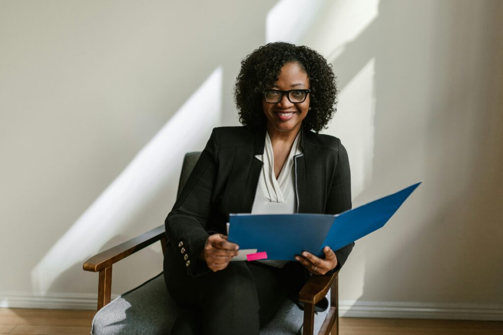 Successful African American woman with glasses and curly hair holding a blue folder indoors.